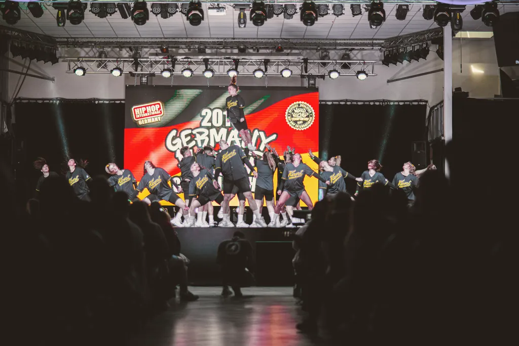 Crew in dark blue outfits building a pyramid on stage, the audience in silhouette in front