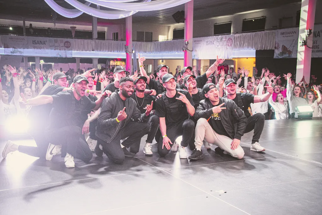 Crew posing on stage, the audience cheering behind them
